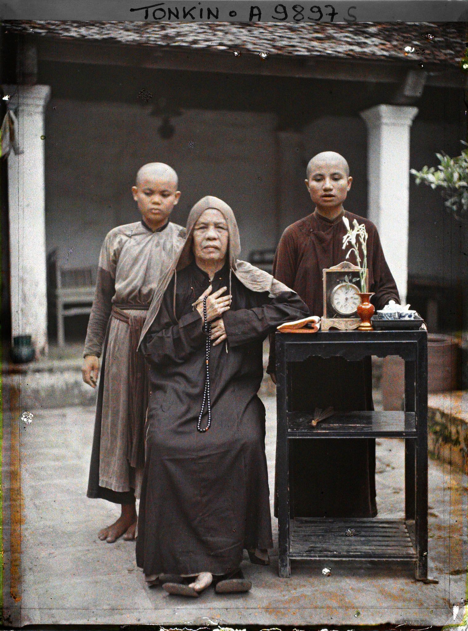Image représentant Une bonzesse et deux novices, près d'une étagère supportant un livre, une pendulette, un vase et un service à thé