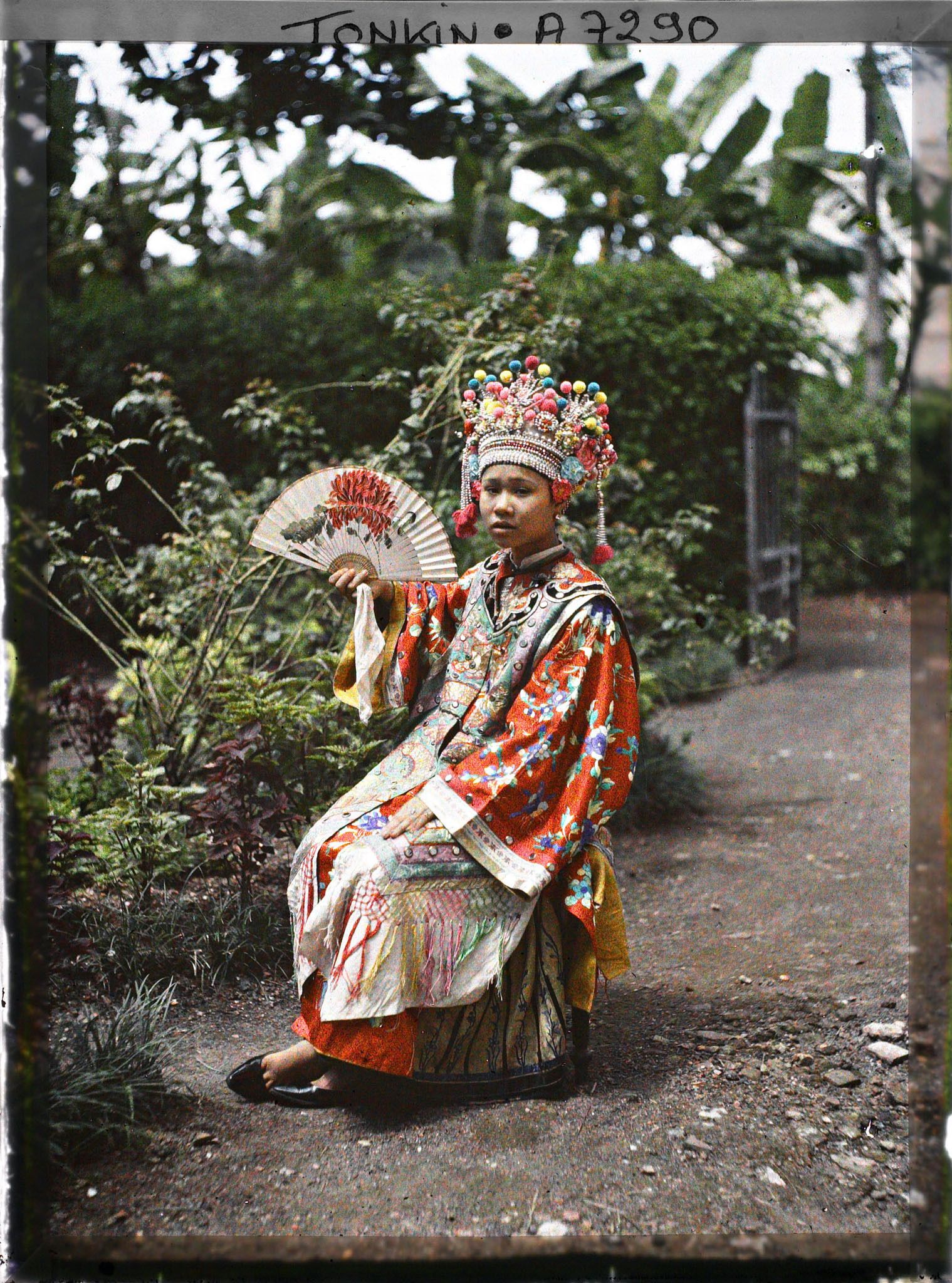 Image représentant Une actrice du Théâtre Saïgonnais, en costume de scène, éventail déployé à la main, assise dans un jardin