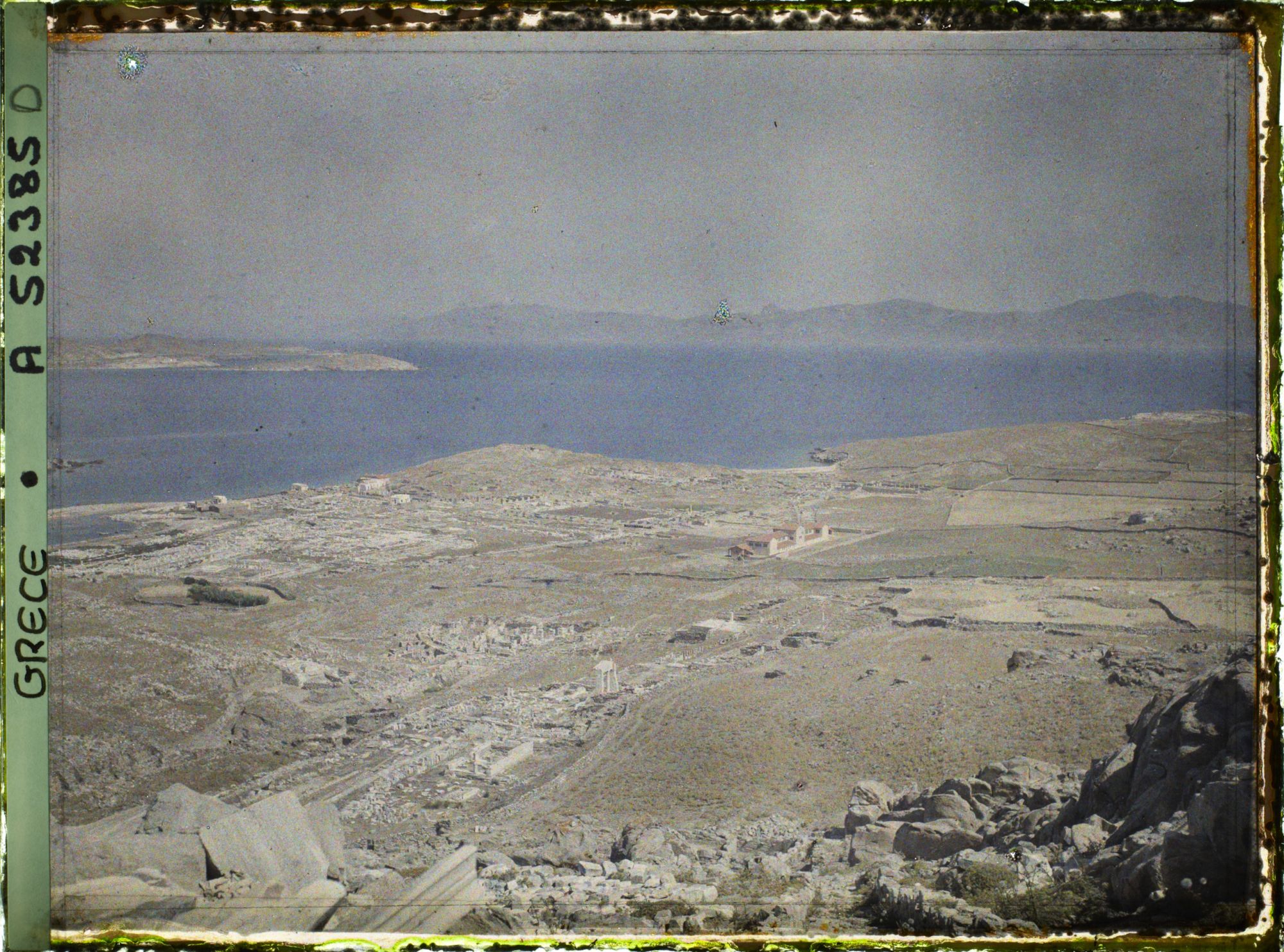 Image représentant Panorama depuis le mont Cynthe en direction du nord-ouest : la ville-haute, puis au bord de la mer, la ville-basse et le port. Au fond, les îles de Syros et plus loin Tinos