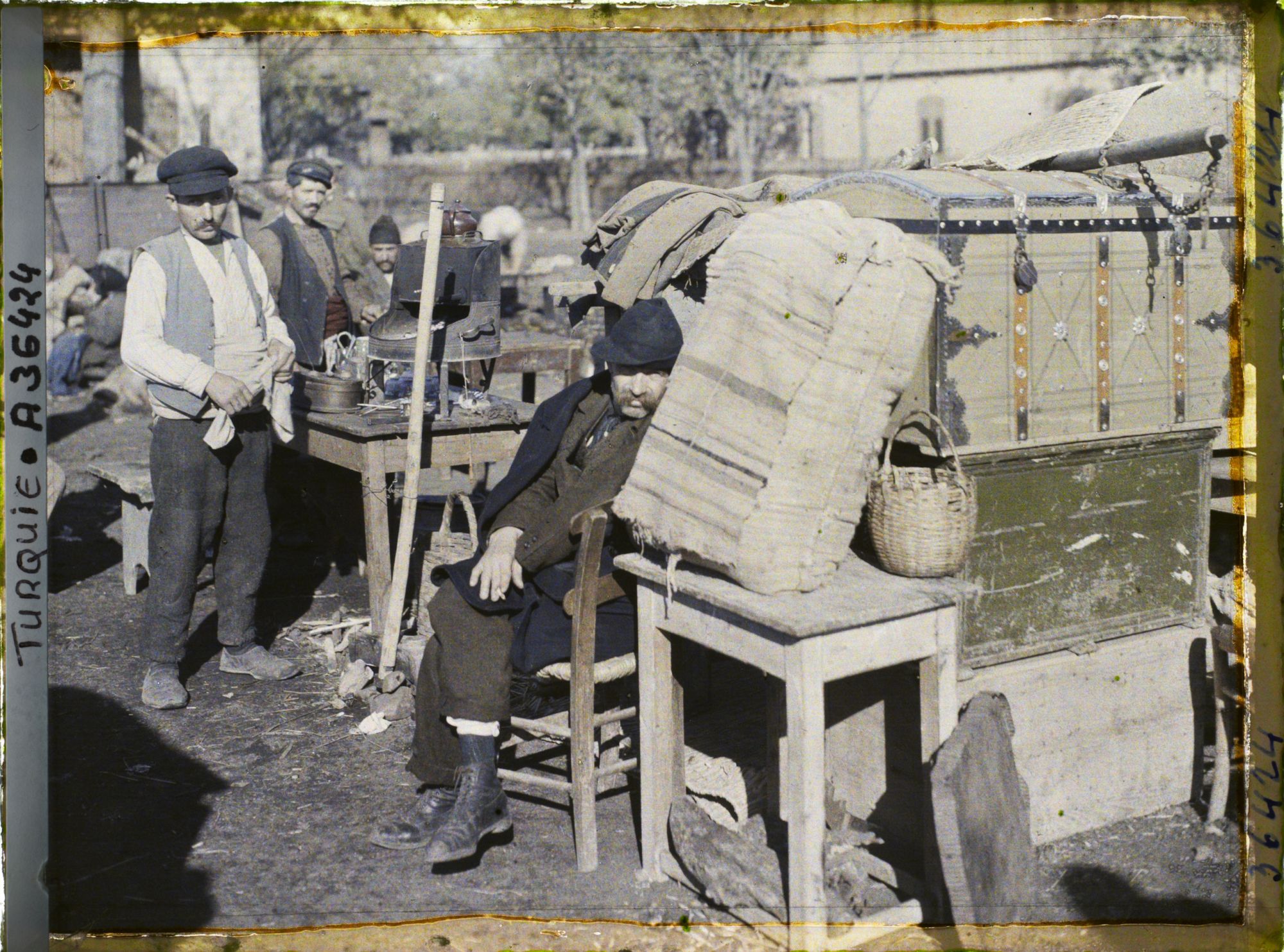 Image représentant Des Grecs s'apprêtant à quitter la ville, improvisant une cuisine en plein air au milieu de caisses et de mobiliers, aux abords de la gare ;