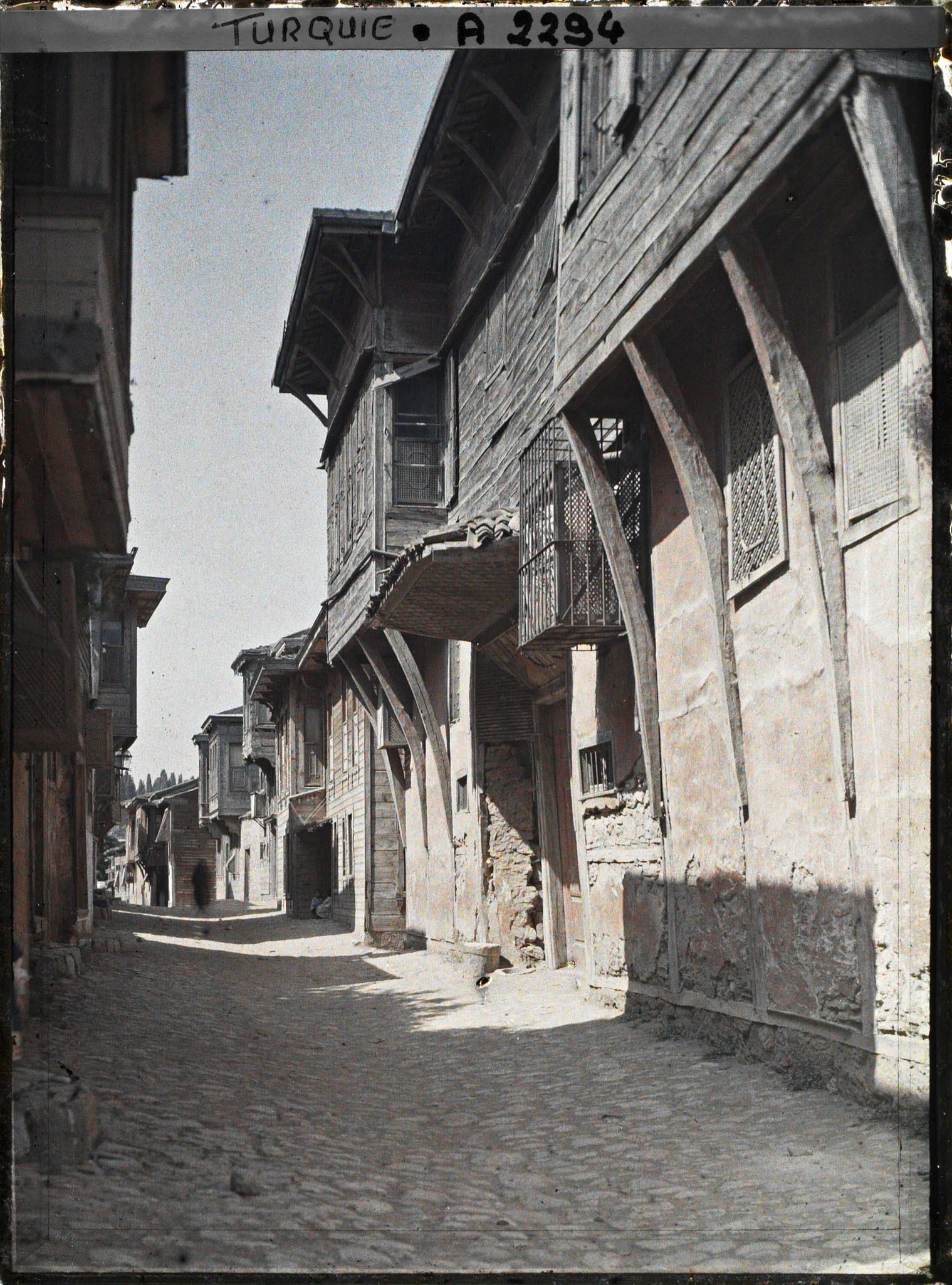 Image représentant Rue pavée, bordée de maisons en dur à moucharabiehs de bois près de Yeni Valide Camii ("mosquée de la Sultane-mère")