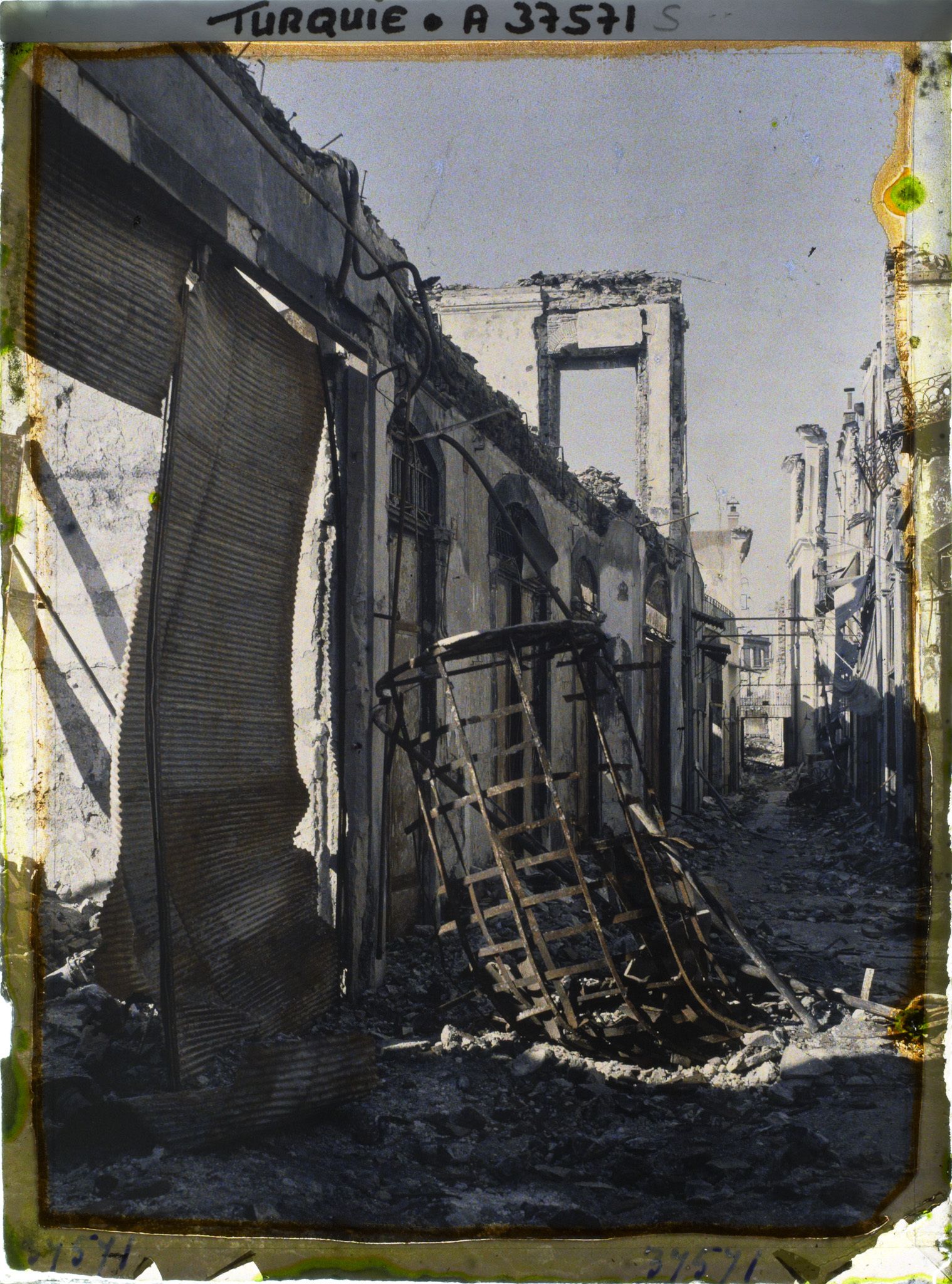 Image représentant Une petite rue en ruines (un rideau de boutique en tôle ondulée dégringolant, une haute cage métallique à terre)
