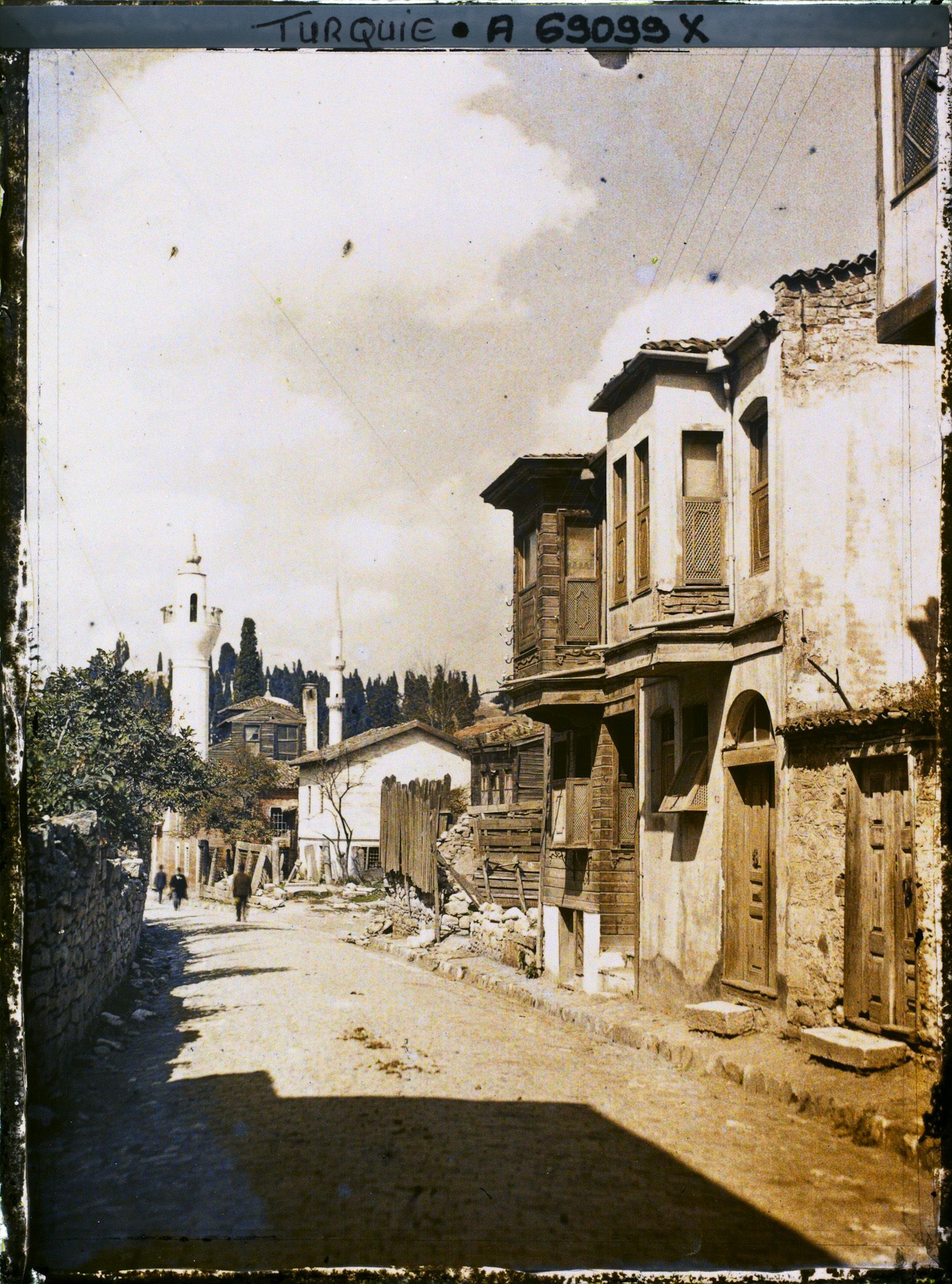 Image représentant Une rue bordée de maisons en dur et en bois,  deux petits minarets à l'arrière plan