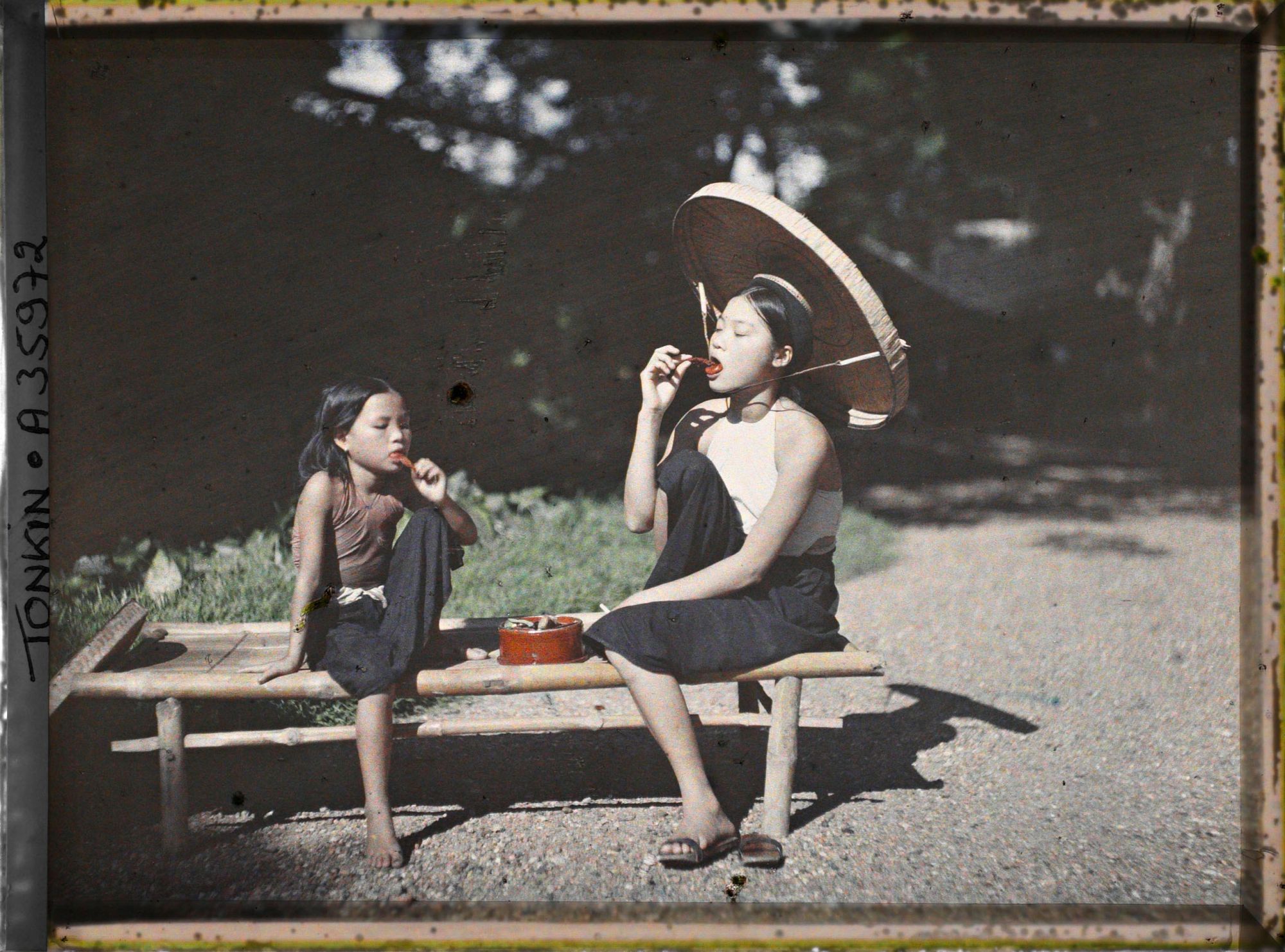 Image représentant Une jeune fille et une fillette chiquant le bétel sont assises sur une banquette en bambou