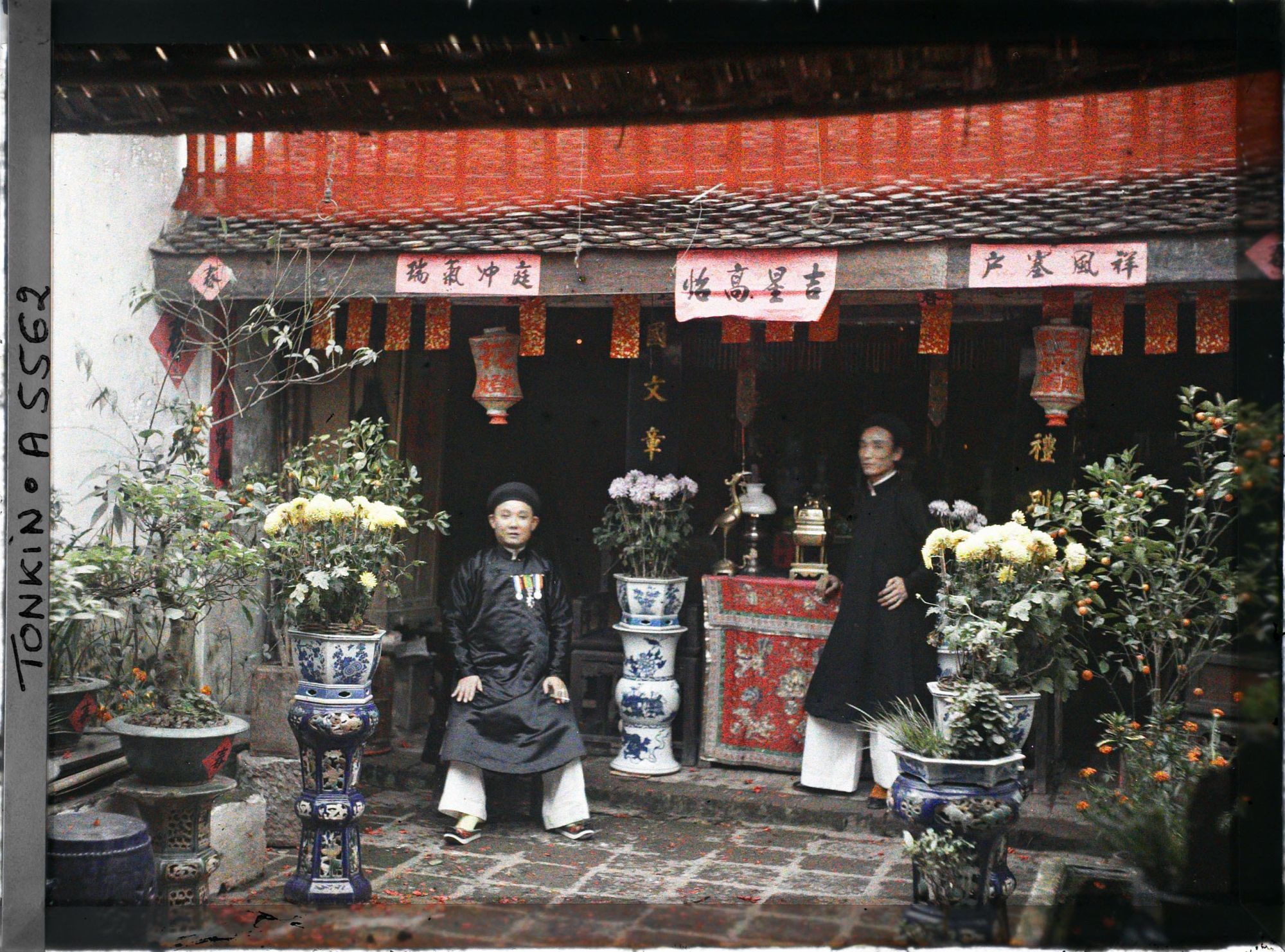 Image représentant Deux notables devant l'autel des ancêtres dans la cour d'une maison aisée, ornée de pots de chrysanthèmes, au moment de la fête du Têt