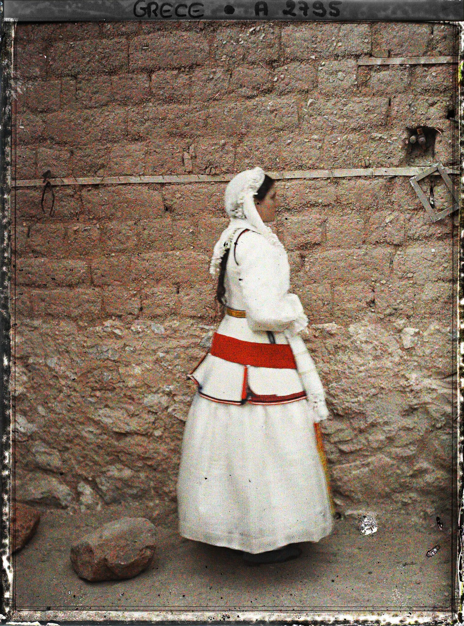 Image représentant Une villageoise en costume de cérémonie (robe blanche, veste à basques, tablier brodé) , de profil