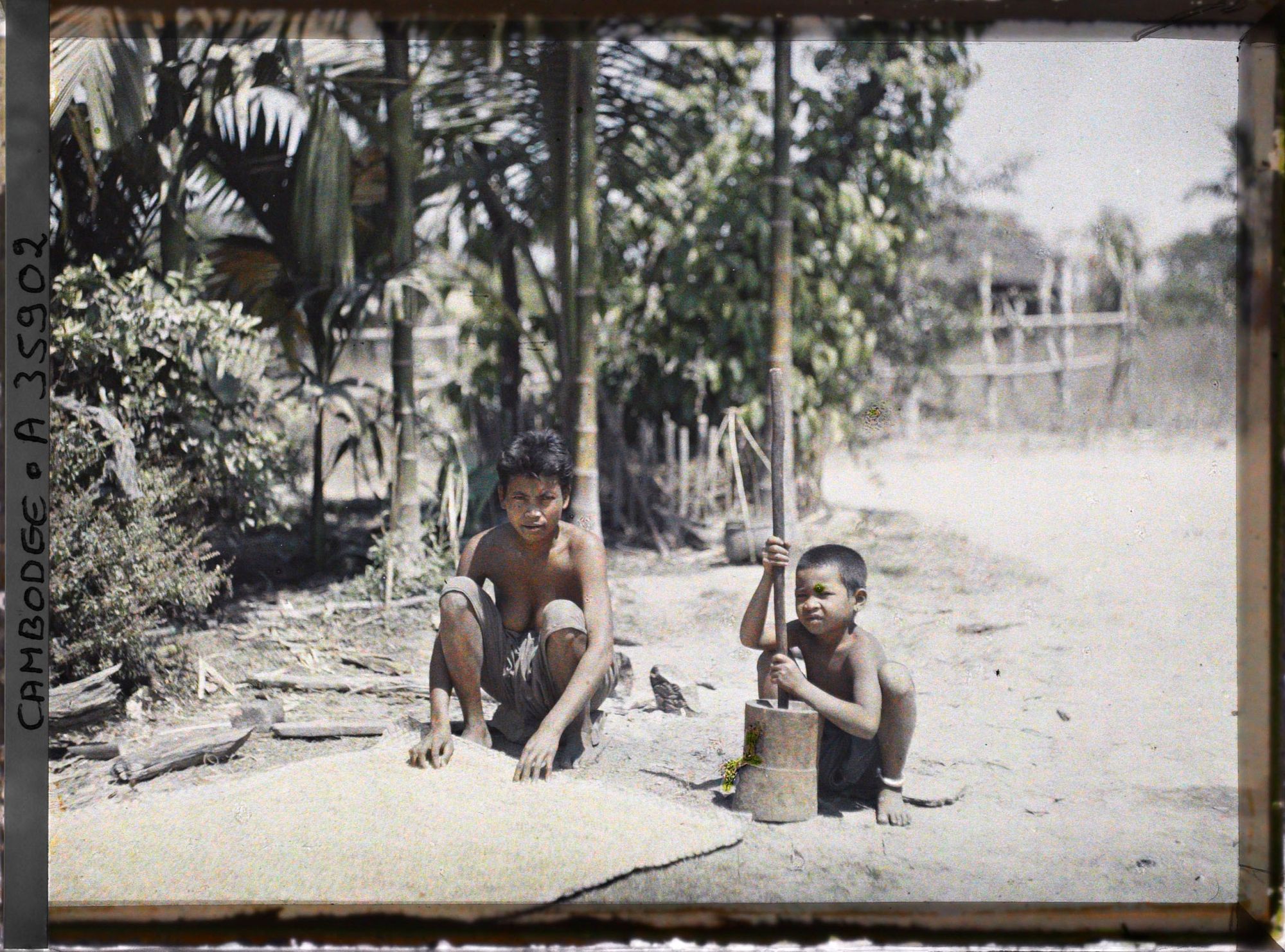 Image représentant Une jeune femme accroupie près d'une natte et un enfant pilonnant du riz