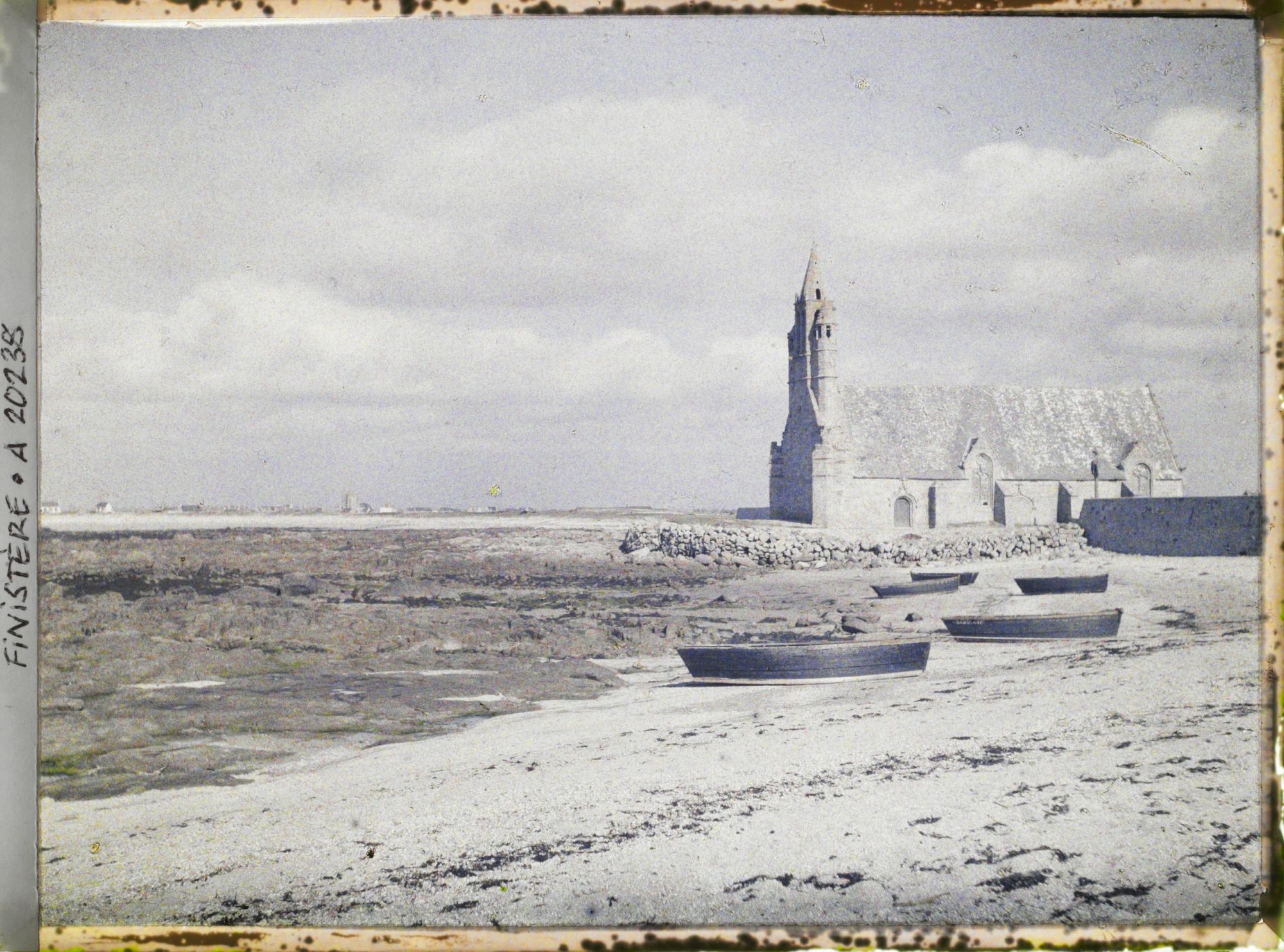Image représentant Barques échouées sur la grève près de la chapelle Notre-Dame-de-la-Joie. A l'horizon, le village de Saint-Guénolé