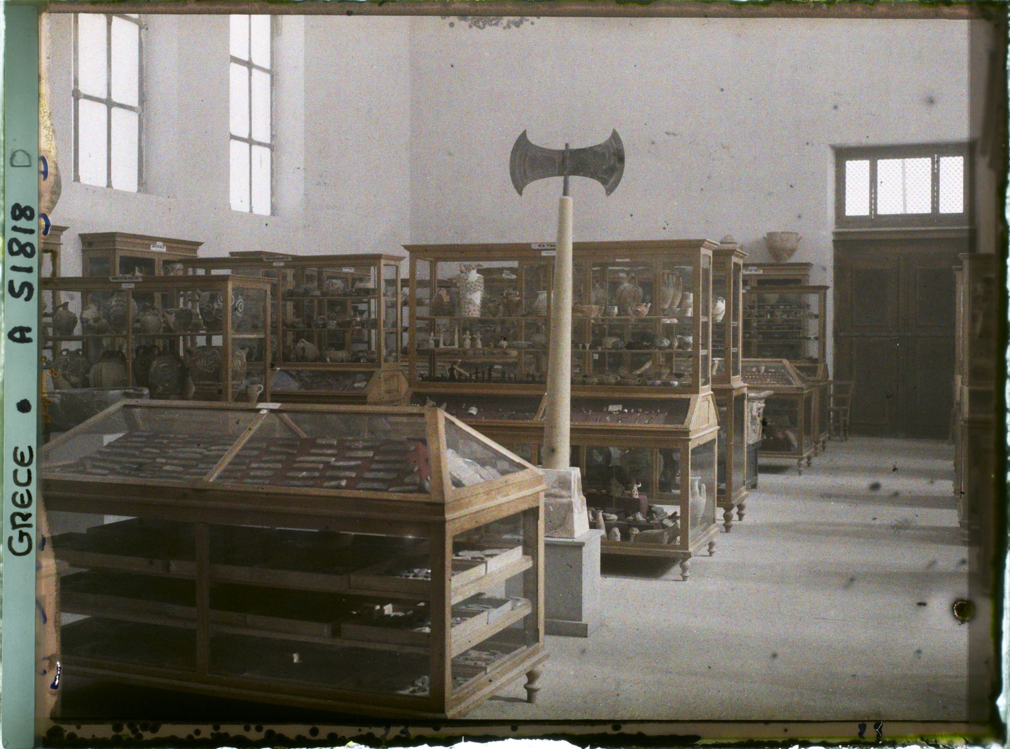 Image représentant Musée archéologique - Vitrines de bois contenant poteries et autres objets. Double hache sur un socle