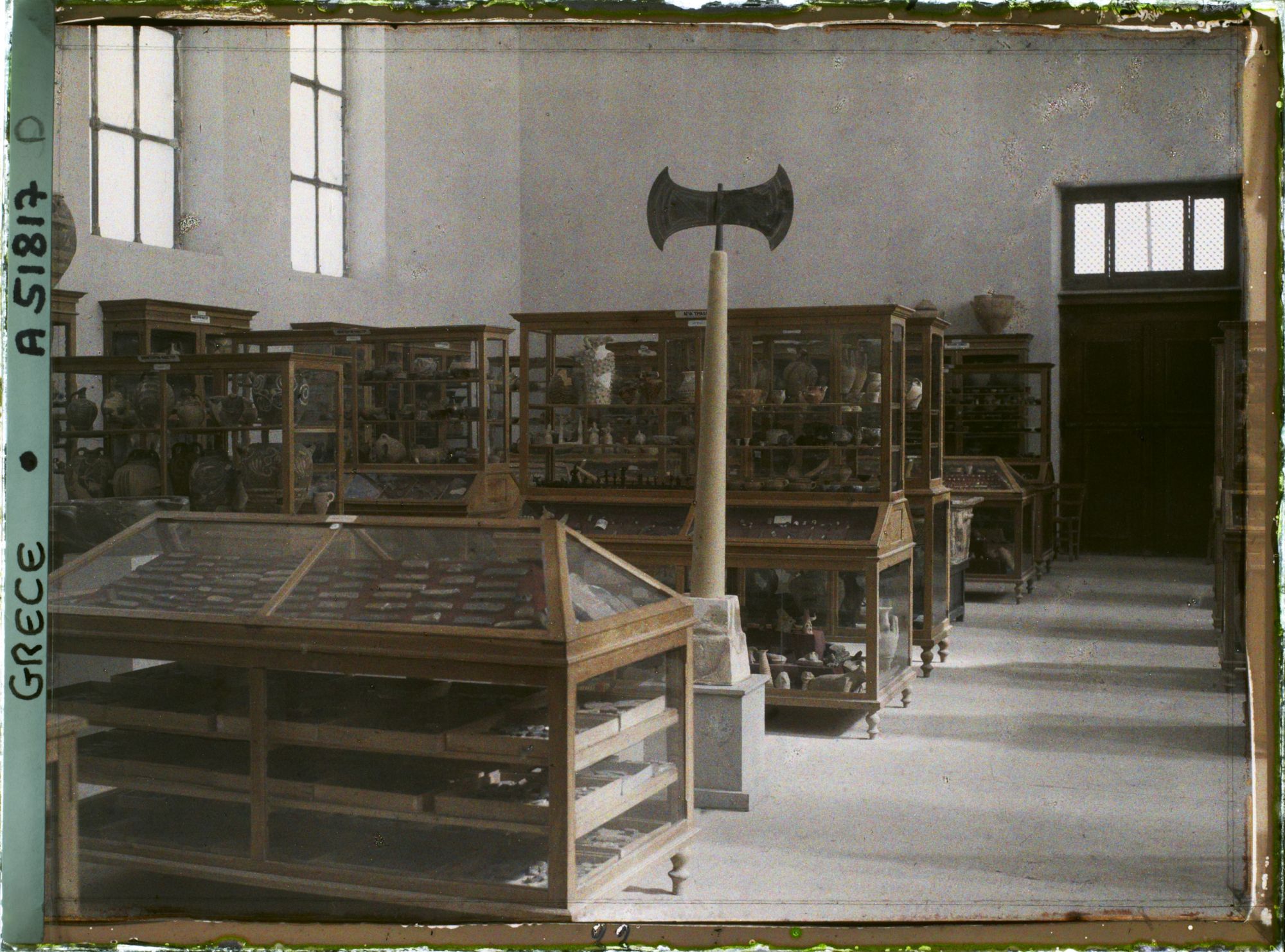 Image représentant Musée archéologique - Vitrines de bois contenant poteries et autres objets. Double hache sur un socle