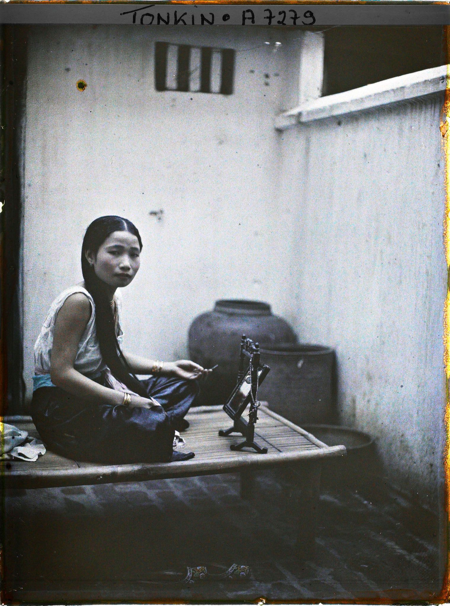 Image représentant Une jeune élégante de classe aisée à sa toilette, assise sur une banquette en bambou dans la cour intérieure de son habitation