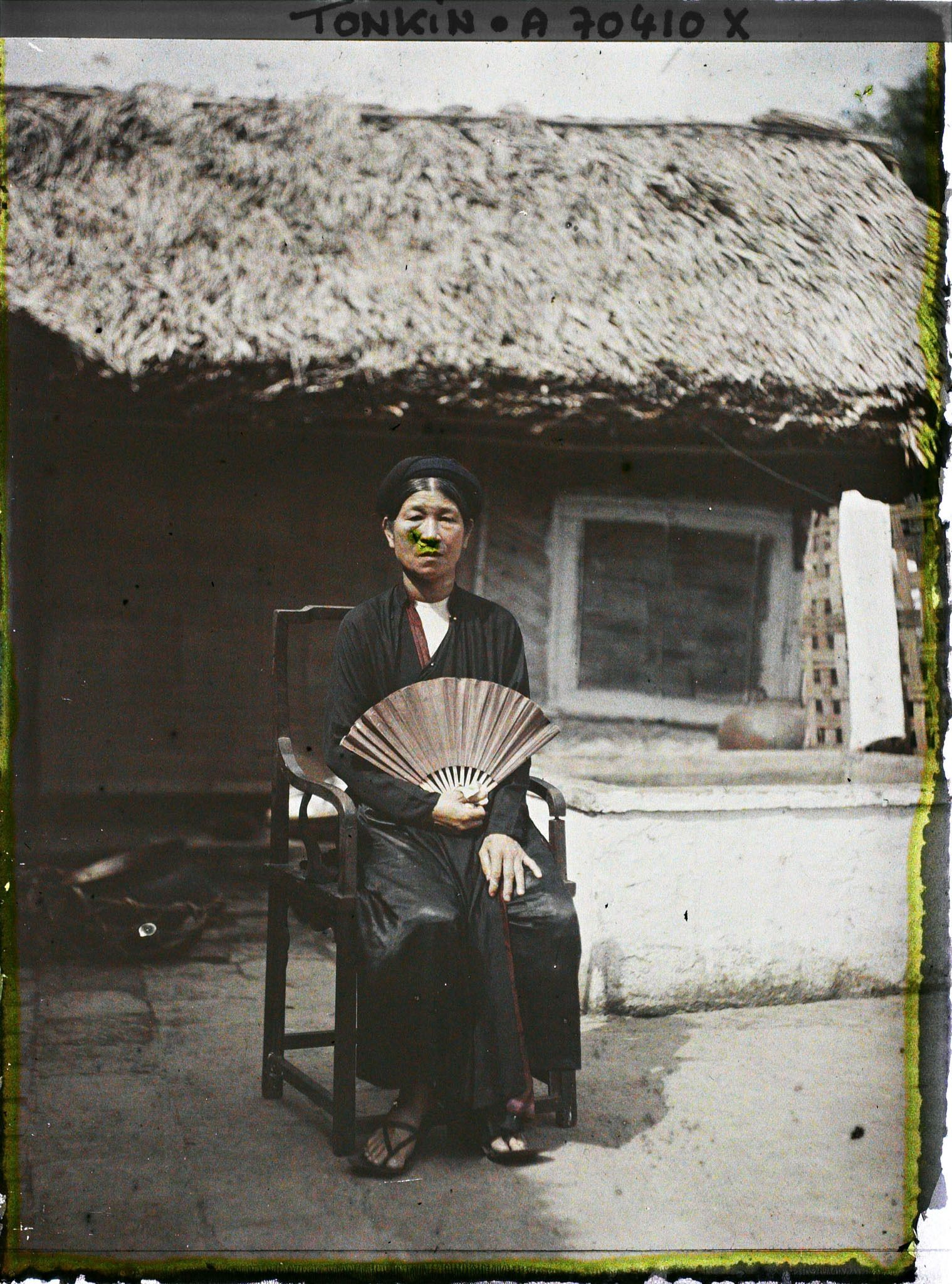 Image représentant La mère de l'un des jeunes époux, assise dans un fauteuil en bois, un éventail déployé à la main, dans une cour intérieure