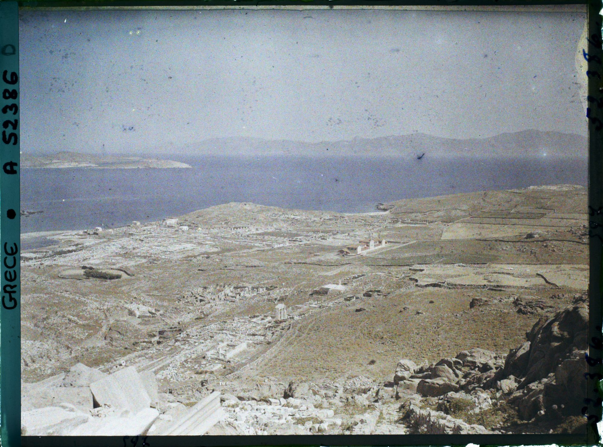 Image représentant Panorama depuis le mont Cynthe en direction du nord-ouest : la ville-haute, puis au bord de la mer, la ville-basse et le port. Au fond, les îles de Syros et plus loin Tinos