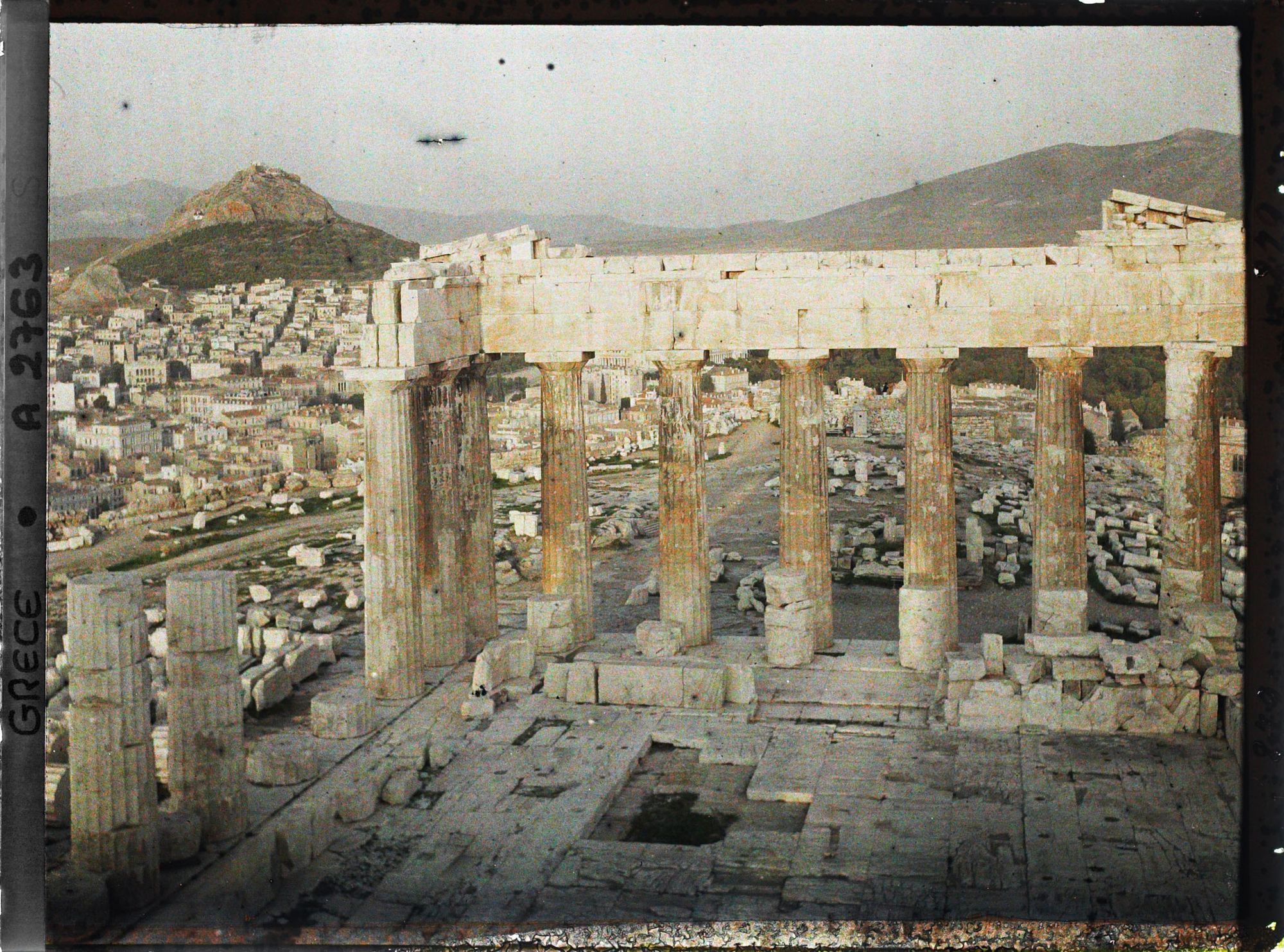 Image représentant Sur l'Acropole, le Parthénon. Vestiges de la colonnade extérieure côté est et derrière eux façade est de l'Acropole, la ville à ses pieds et le Mont Lycabette (vue plongeante)