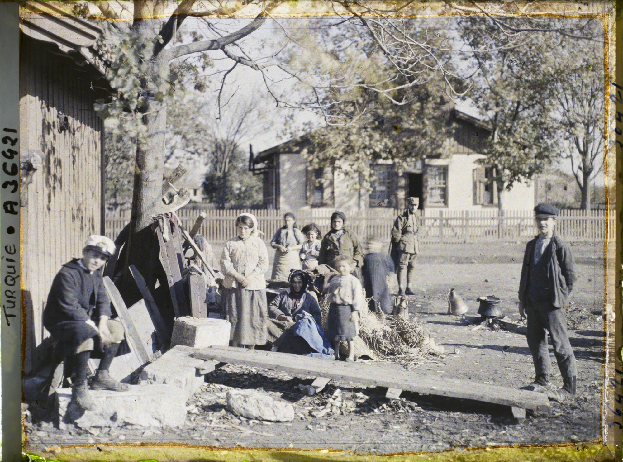 Image représentant Femmes et enfants grecs s'apprêtant à quitter la ville, aux abords de la gare ; derrière eux, un soldat (grec ou allié ?) ; près du groupe, un foyer avec une marmite et un récipent en cuivre