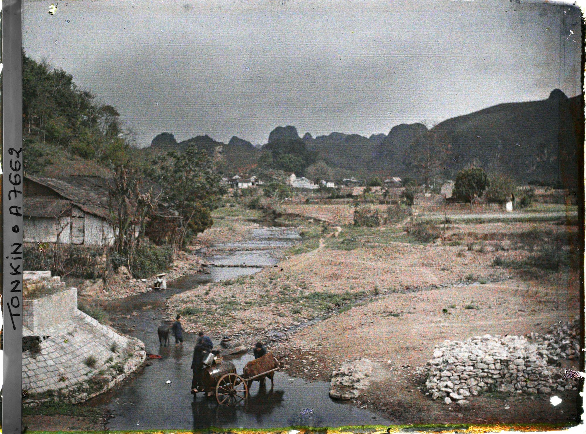Image représentant Le village et la rivière, vus du pont, au pied duquel une charrette munie d'un tonneau s'approvisionne en eau