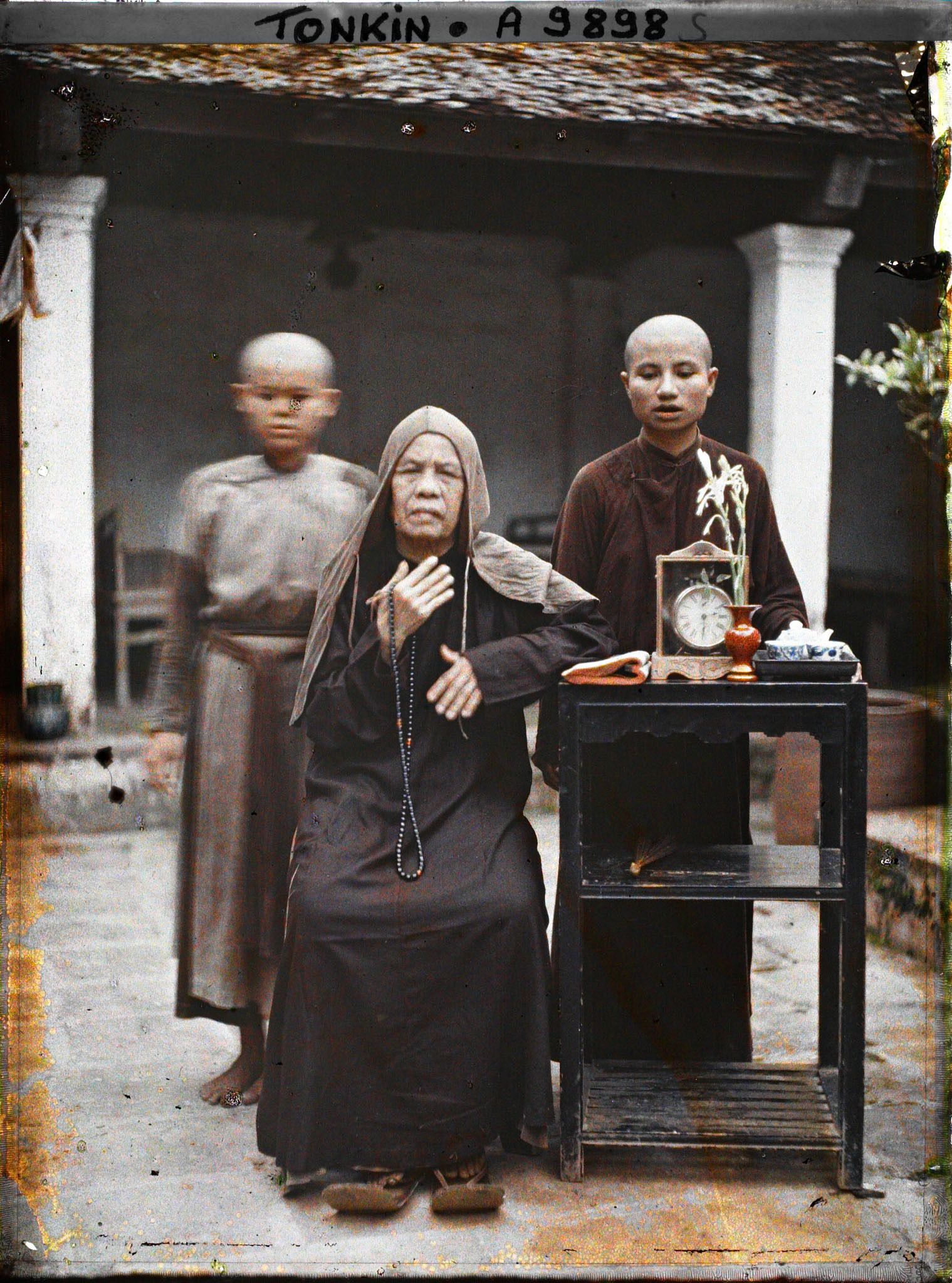 Image représentant Une bonzesse et deux novices, près d'une étagère supportant un livre, une pendulette, un vase et un service à thé