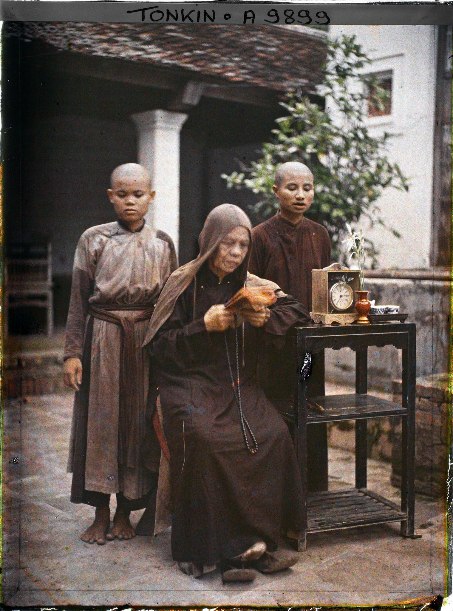 Image représentant Une bonzesse lisant devant deux novices, accoudée à une étagère supportant une pendulette, un vase et un service à thé