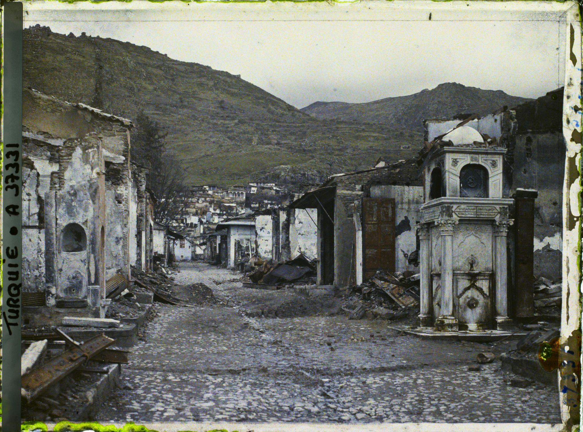 Image représentant Une rue bordée de bâtiments endommagés. A droite, une fontaine