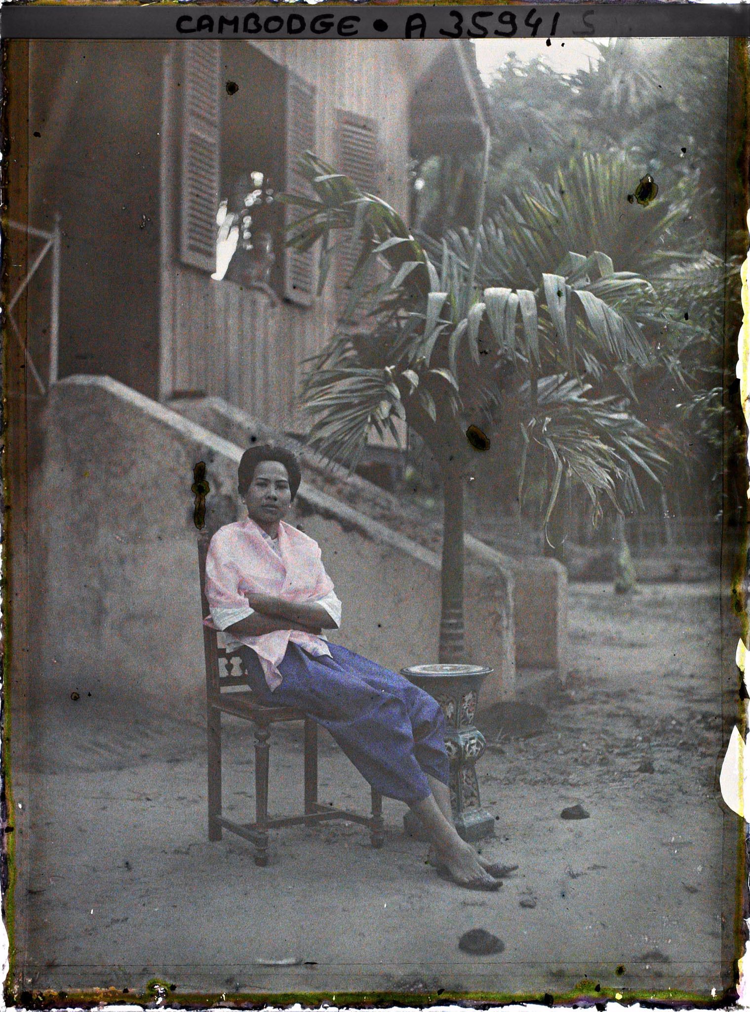 Image représentant Femme en sampot violet, assise sur une chaise de bois tournée devant le perron d'une maison