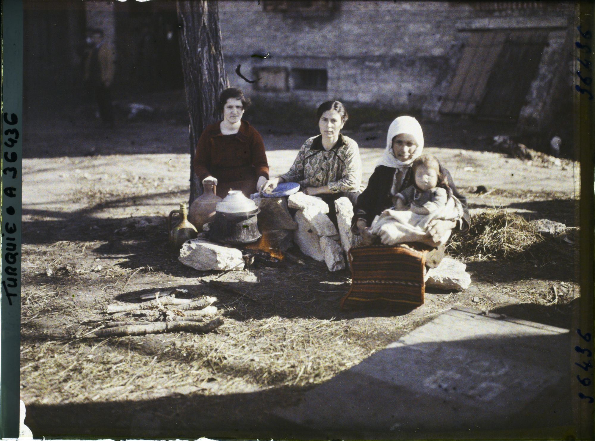 Image représentant Trois femmes grecques dont l'une tenant un enfant, s'apprêtant à quitter la ville, assises aux abords de la gare, près d'un foyer improvisé