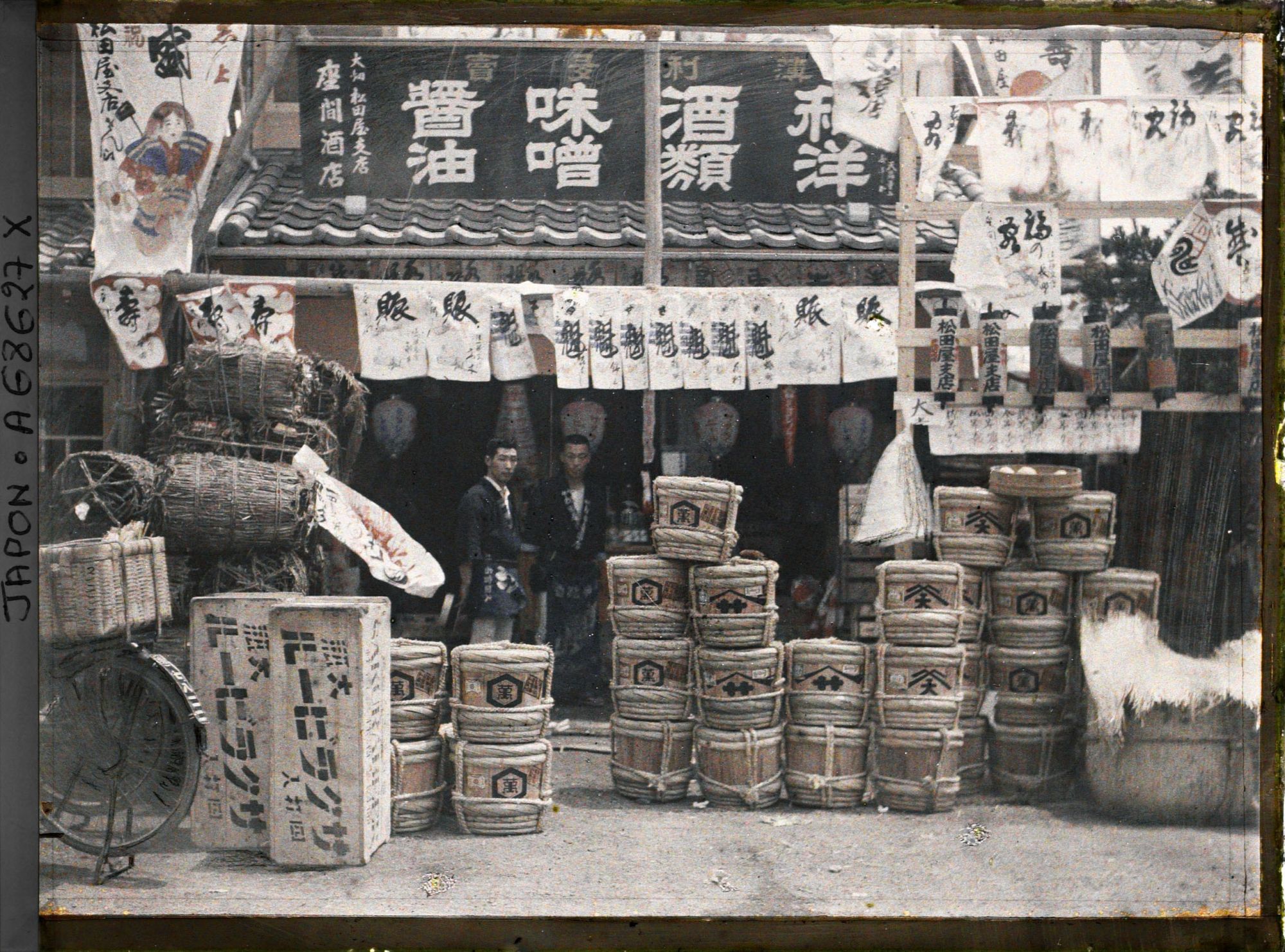 Image représentant Epiciers à l'entrée de leur boutique, devant laquelle sont exposés des fûts de bière, de "miso" (pâte de haricots de soja fermentée), de saké et du charbon de bois
