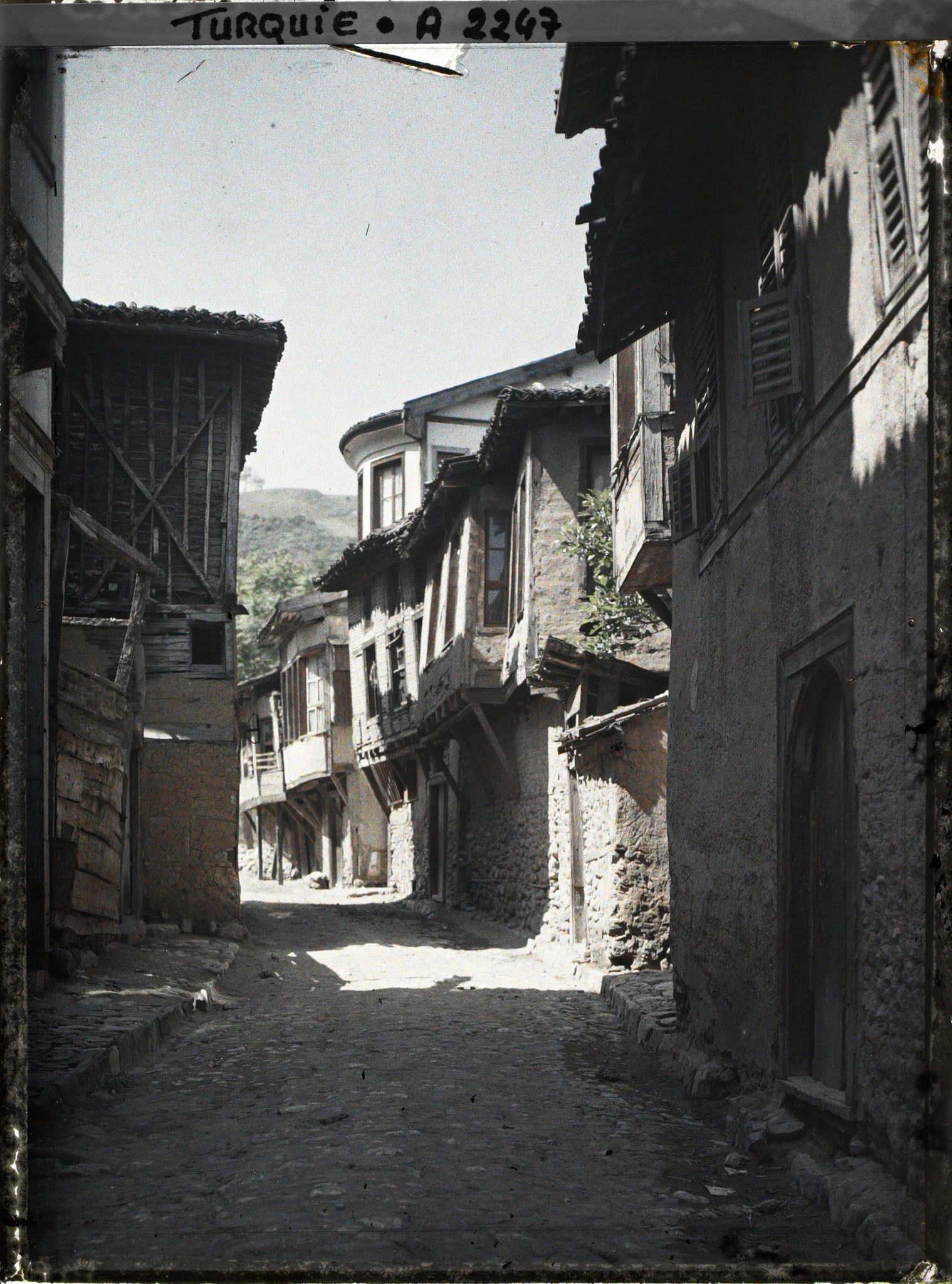 Image représentant Rue étroite pavée, bordée de maisons. Celles du fond à droite sont à encorbellement