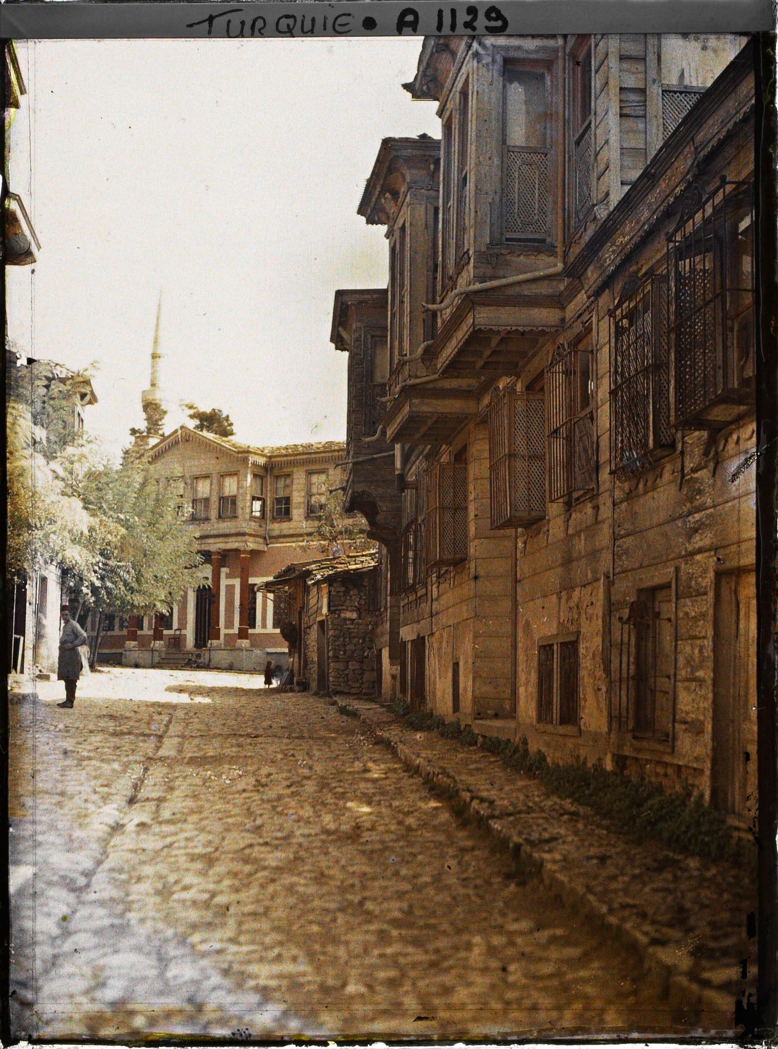 Image représentant Une rue pavée d'Eyüp bordée de maisons à étages de bois en saillie (au fond, un minaret)