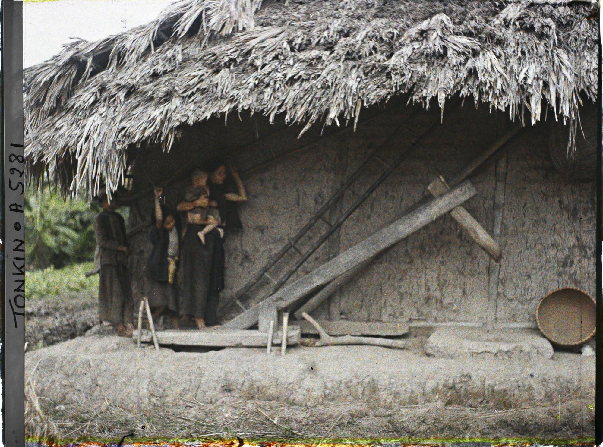Image représentant Une femme accompagnée d'enfants actionnant au pied le pilon à blanchir le riz, dans une cour de ferme après la moisson du riz