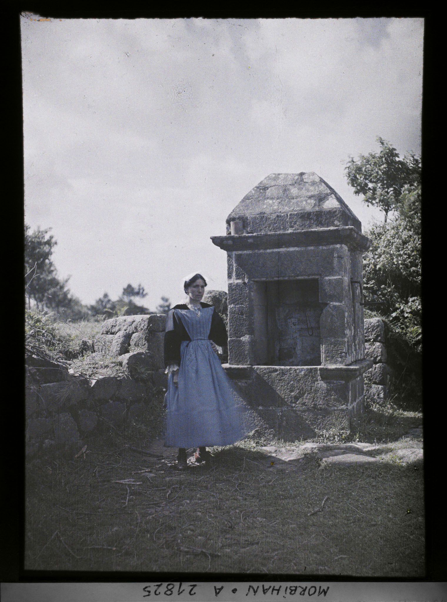 Image représentant Une Bretonne en costume traditionnel posant devant un puits, sur la route d'Auray à Carnac