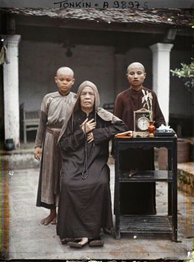 Image représentant Une bonzesse et deux novices, près d'une étagère supportant un livre, une pendulette, un vase et un service à thé