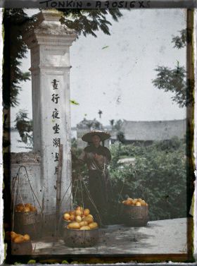 Image représentant Une femme annamite entourée de paniers de fruits (ou légumes), pose pour le photographe près d'un portique au temple Ngoc-so'n