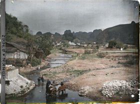 Image représentant Le village et la rivière, vus du pont, au pied duquel une charrette munie d'un tonneau s'approvisionne en eau