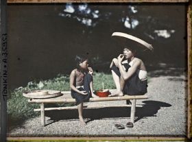 Image représentant Une jeune fille et une fillette chiquant le bétel sont assises sur une banquette en bambou