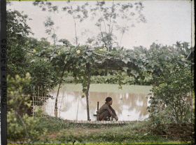 Image représentant Un homme accroupi au bord d'un étang, à l'ombre d'une pergola