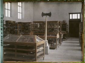 Image représentant Musée archéologique - Vitrines de bois contenant poteries et autres objets. Double hache sur un socle