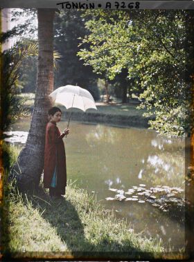 Image représentant Une jeune femme de classe aisée, s'abritant sous une ombrelle blanche au bord d'une pièce d'eau dans un jardin public