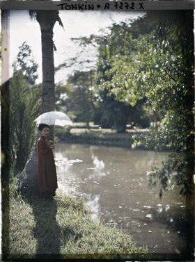 Image représentant Une jeune femme de classe aisée, s'abritant sous une ombrelle blanche au bord d'une pièce d'eau dans un jardin public