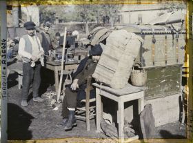 Image représentant Des Grecs s'apprêtant à quitter la ville, improvisant une cuisine en plein air au milieu de caisses et de mobiliers, aux abords de la gare ;