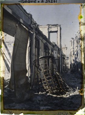 Image représentant Une petite rue en ruines (un rideau de boutique en tôle ondulée dégringolant, une haute cage métallique à terre)