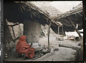 Image représentant Une femme actionnant une meule à farine sous l'auvent d'une maison