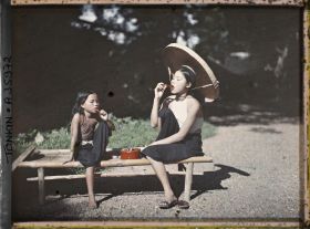 Image représentant Une jeune fille et une fillette chiquant le bétel sont assises sur une banquette en bambou