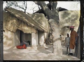 Image représentant Une femme assise sous l'auvent d'une maison ; au premier plan à droite, un jeune homme et un enfant