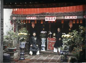 Image représentant Deux notables devant l'autel des ancêtres dans la cour d'une maison aisée, ornée de pots de chrysanthèmes, au moment de la fête du Têt