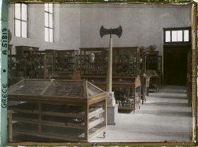 Image représentant Musée archéologique - Vitrines de bois contenant poteries et autres objets. Double hache sur un socle