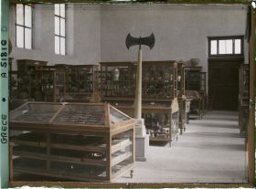 Image représentant Musée archéologique - Vitrines de bois contenant poteries et autres objets. Double hache sur un socle
