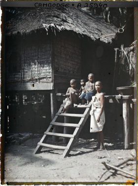 Image représentant Une femme en deuil et des enfants près de l'échelle permettant d'accèder à une maison sur pilotis