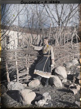 Image représentant Jeune fille, en robe brodée, devant un mur de petit bois avec à l'arrière-plan l'église