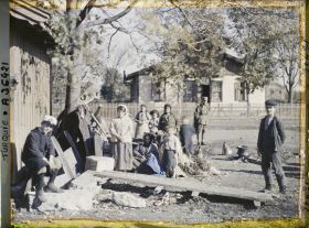 Image représentant Femmes et enfants grecs s'apprêtant à quitter la ville, aux abords de la gare ; derrière eux, un soldat (grec ou allié ?) ; près du groupe, un foyer avec une marmite et un récipent en cuivre