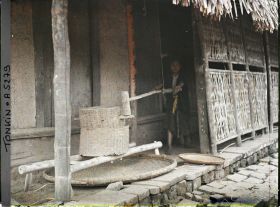 Image représentant Une jeune fille actionnant un moulin à décortiquer le paddy, dans une cour de ferme après la moisson du riz